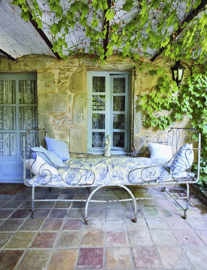 Kunstvolles Metallbett im Shabby Stil auf einer überdachten Terrasse mit rankendem Wein und Terrakottafliesen