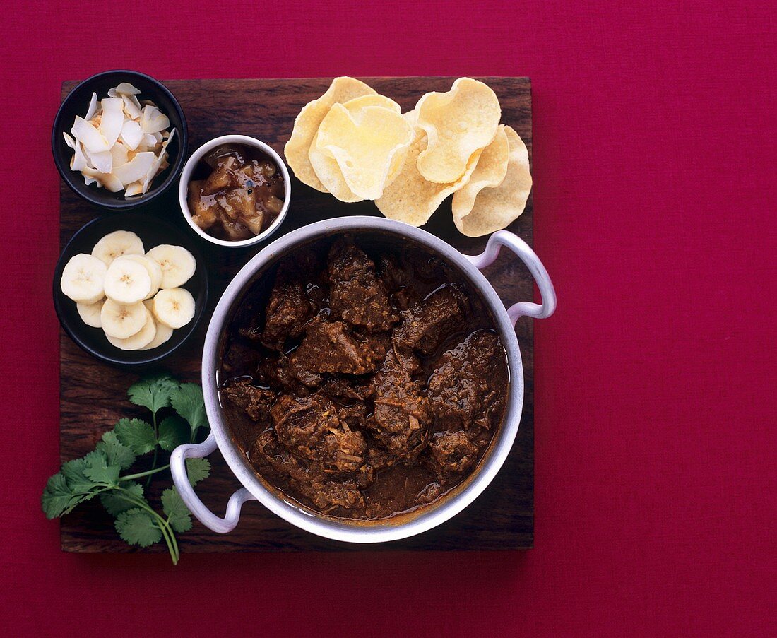 Beef curry in pot with mini-poppadoms and accompaniments