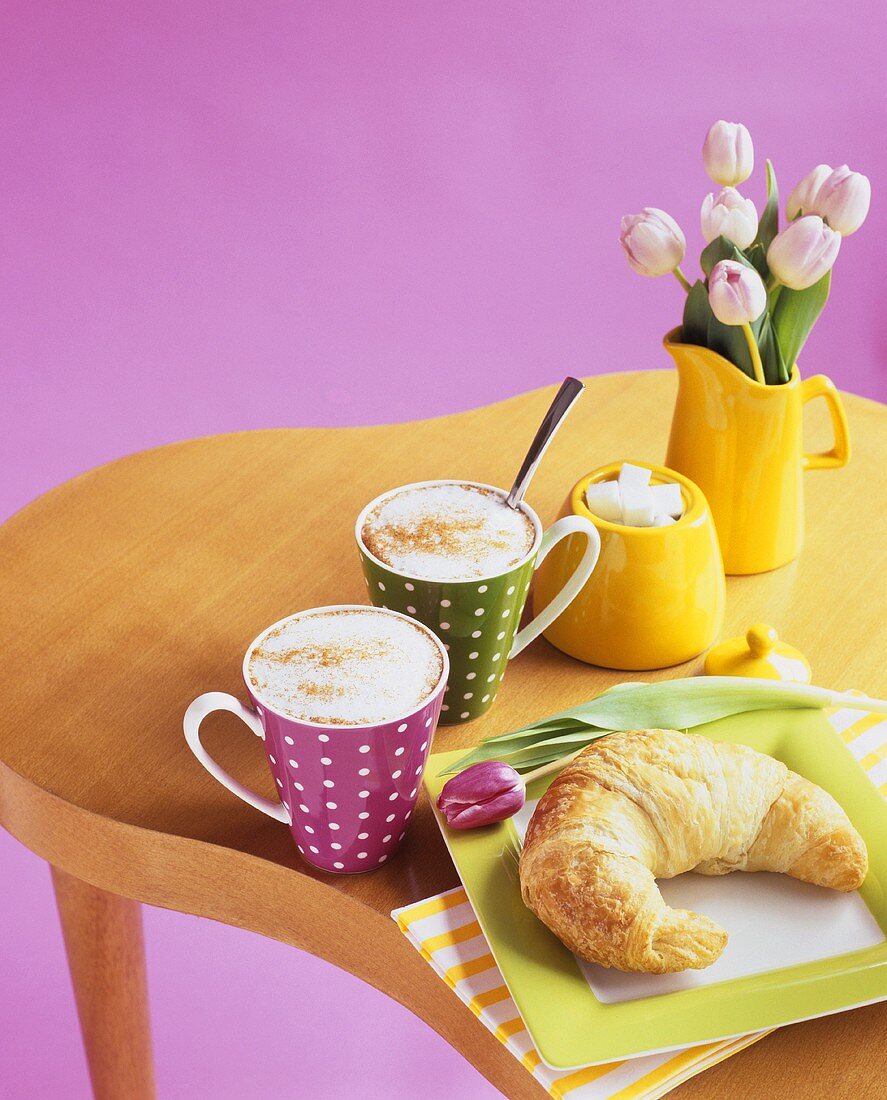 Croissant mit zwei Tassen Cappuccino und Tulpen in der Vase