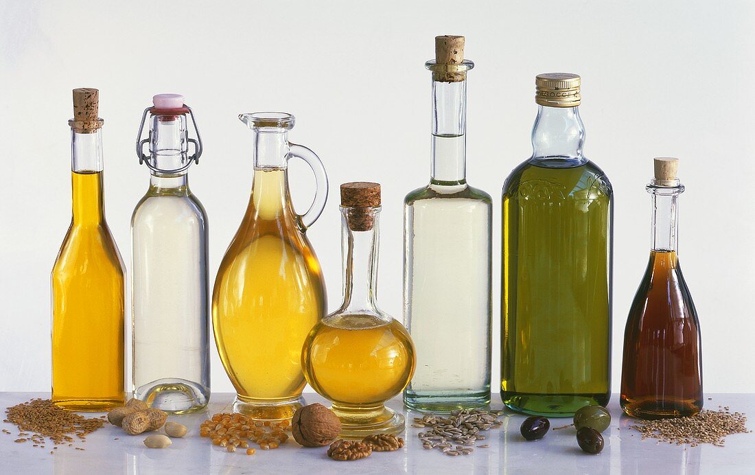Sieben verschiedene Öle in Flaschen, zugehörige Rohstoffe
