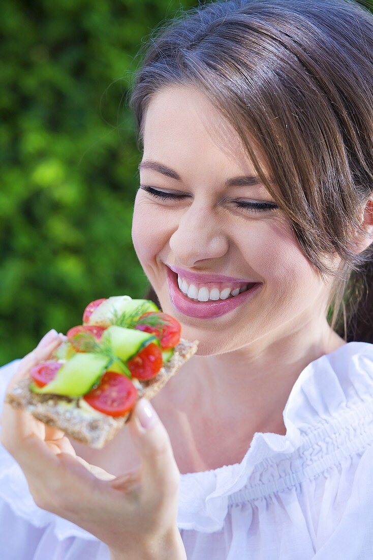 Young woman eating crispbread topped with tomato & cucumber