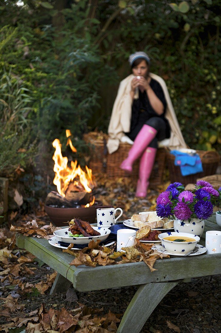 Junge Frau sitzt am Lagerfeuer mit herbstlich gedecktem Tisch