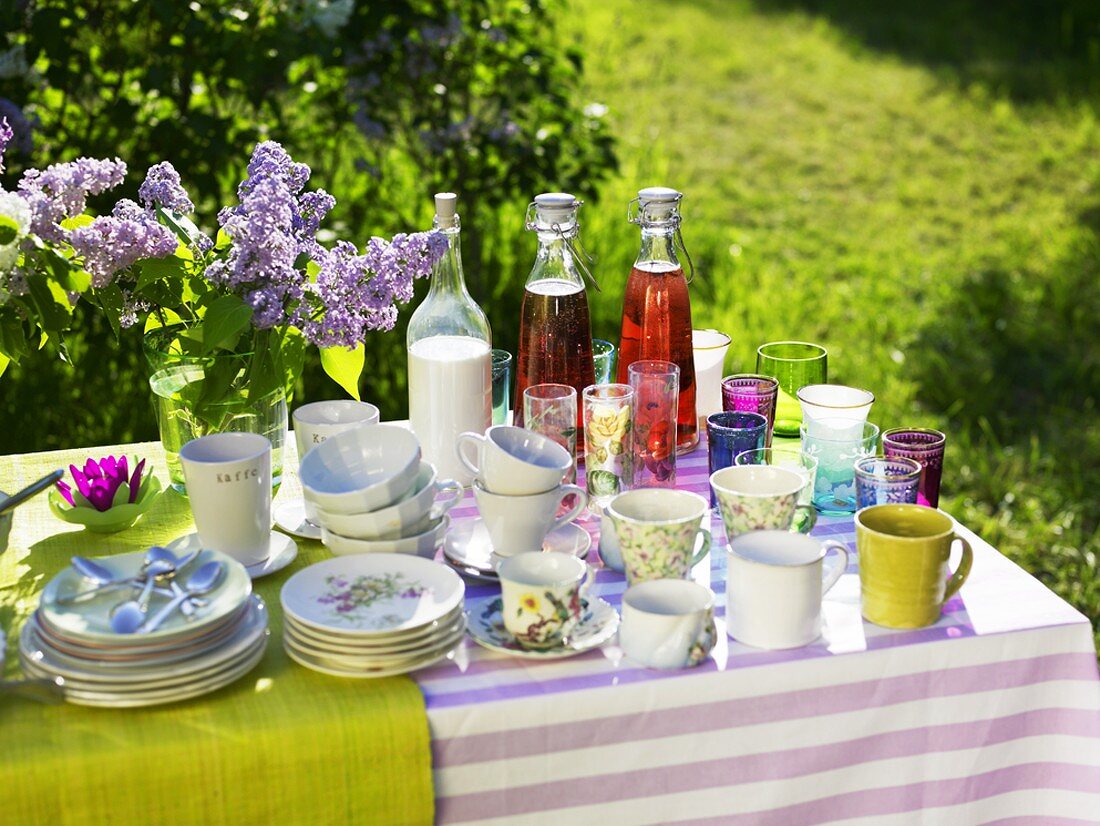 Tisch mit Tassen, Gläsern, Getränken & lila Flieder im Freien