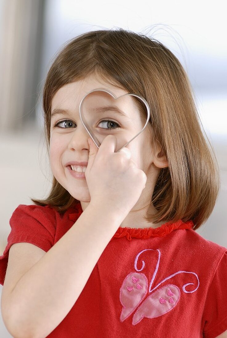 Girl holding heart-shaped biscuit cutter in front of her eye