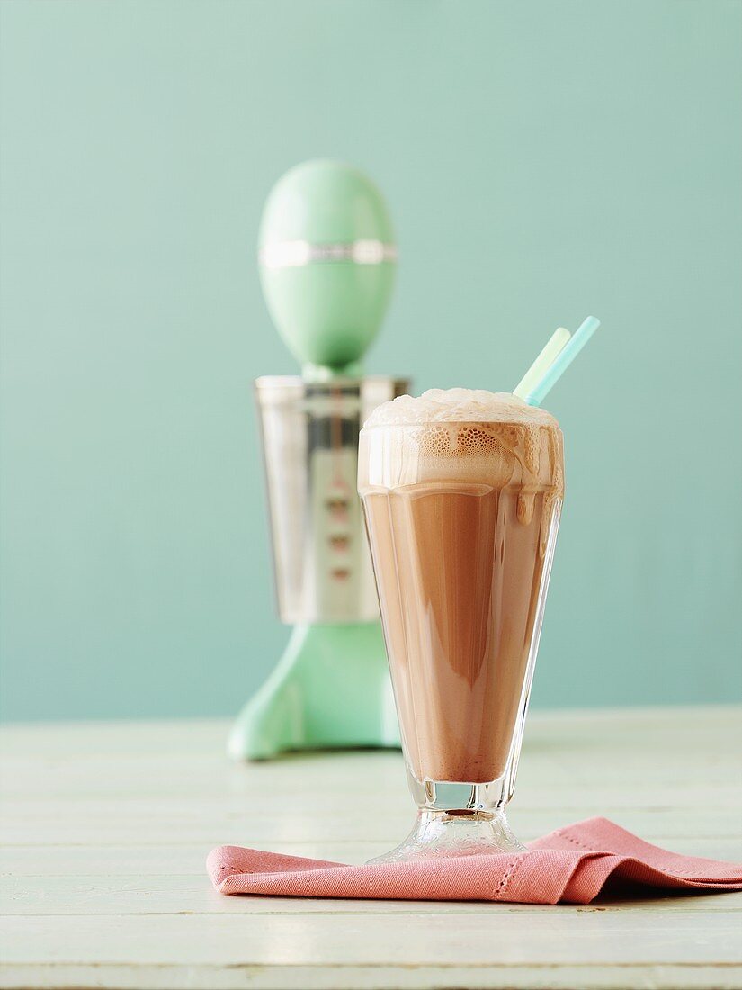 Ein Glas Schokoladenmilchshake, im Hintergrund ein Mixer