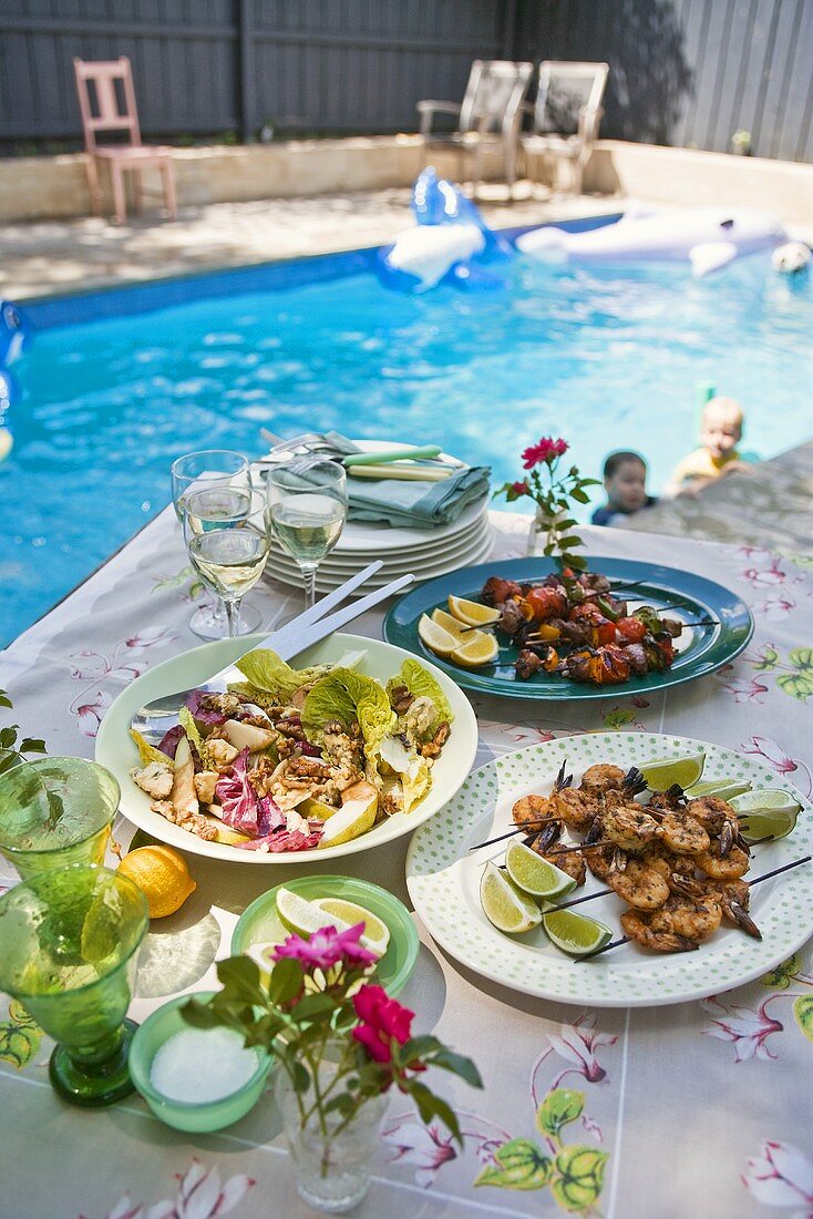 Verschiedene Grillspiesse und Salat auf einem Tisch am Pool