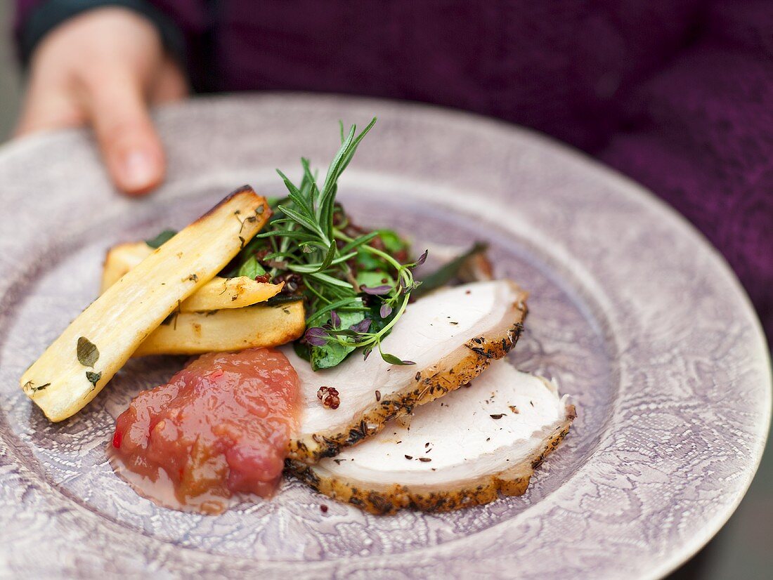 A hand holding a plate of sliced meat, apple chutney and parsnips