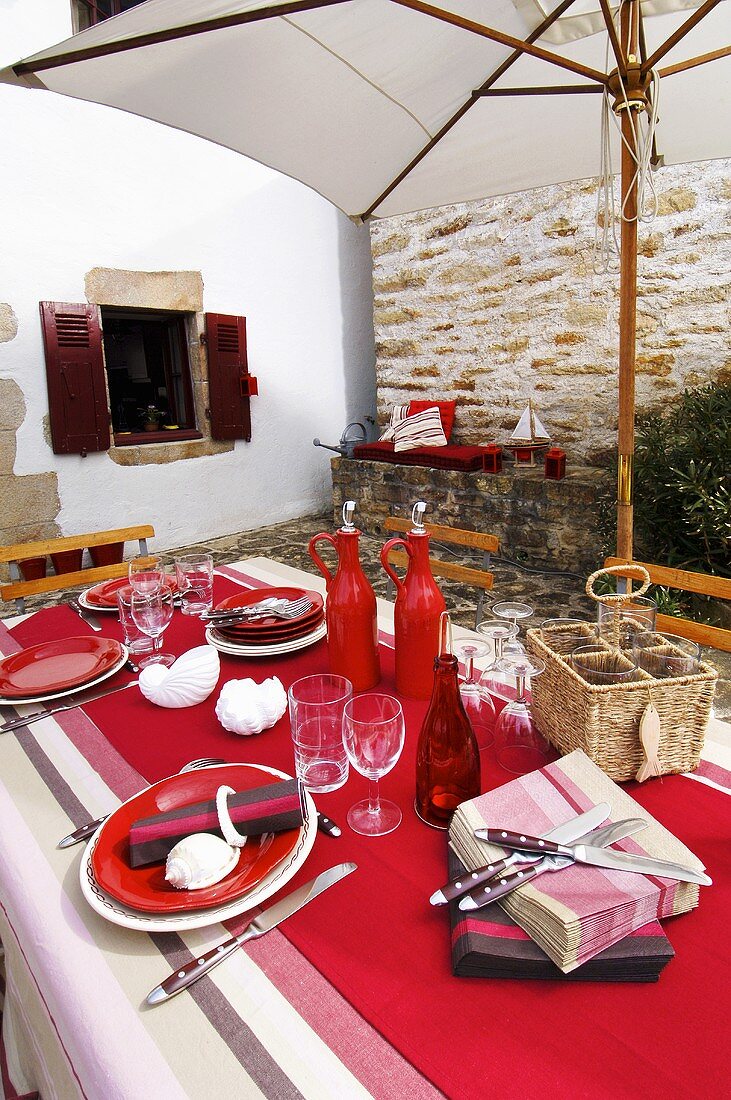 Gedeckter Tisch in Rot mit maritimer Deko auf der Terrasse unter weißem Sonnenschirm