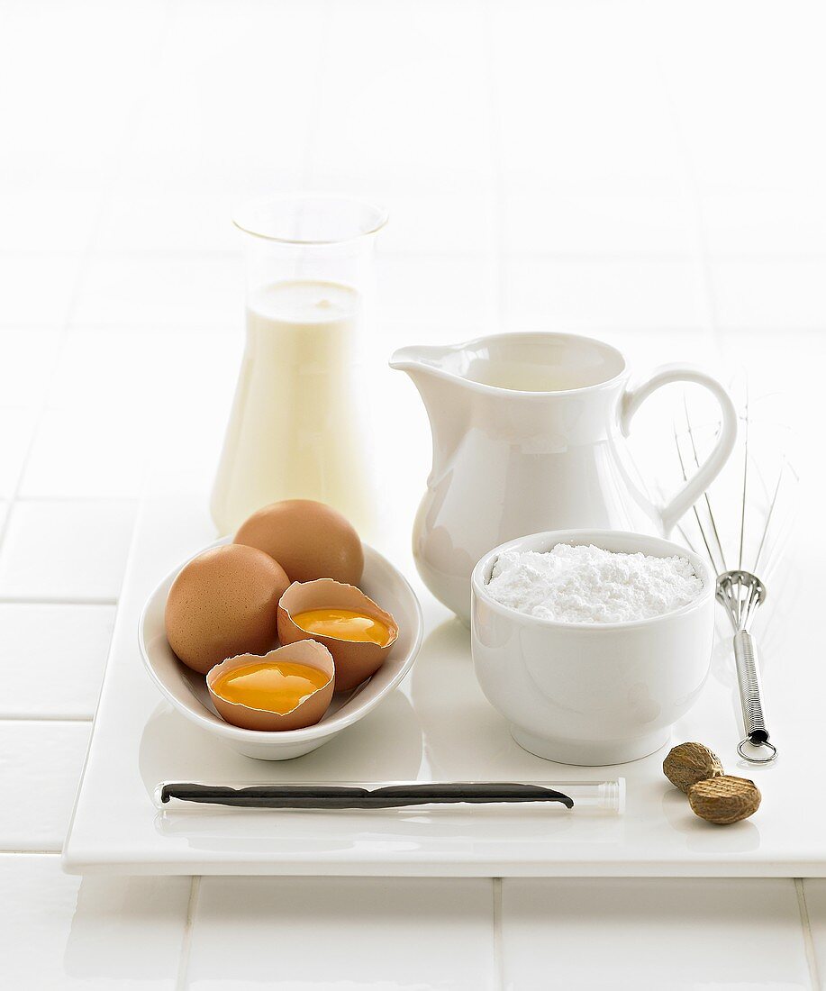Verschiedene Backzutaten (Eier, Zucker, Vanilleschote, Muskatnuss, Milch) und Schneebesen