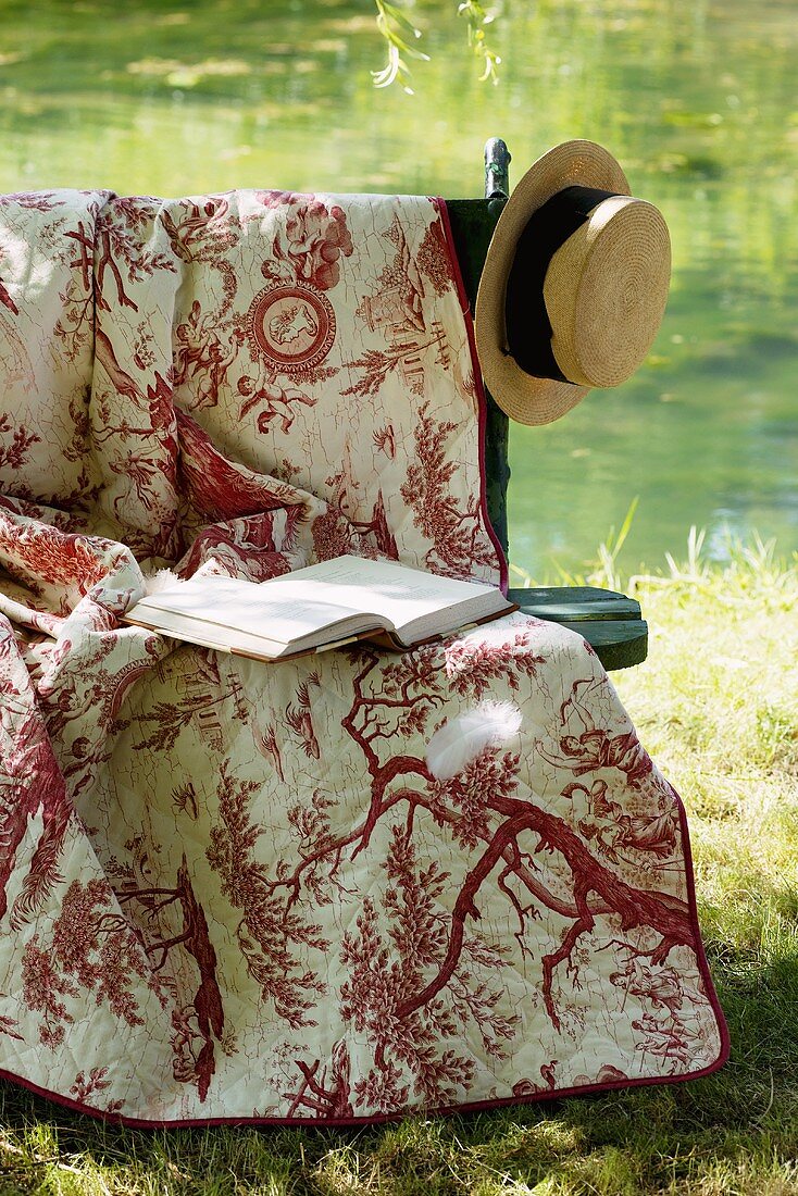 Romantic garden seat with cover, book and straw hat by pond