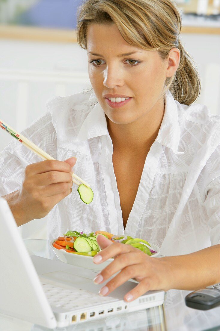 Frau isst Gemüsesalat mit Essstäbchen vor Laptop
