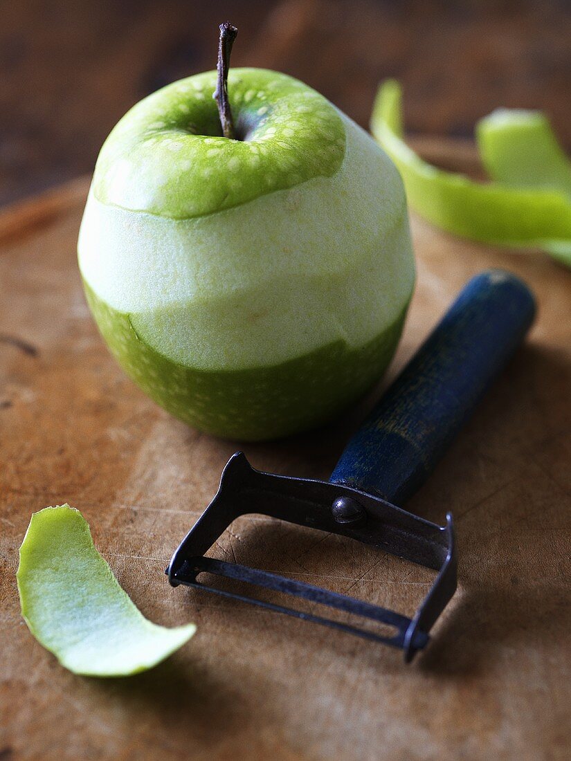 Half-peeled green apple with peeler on wooden background