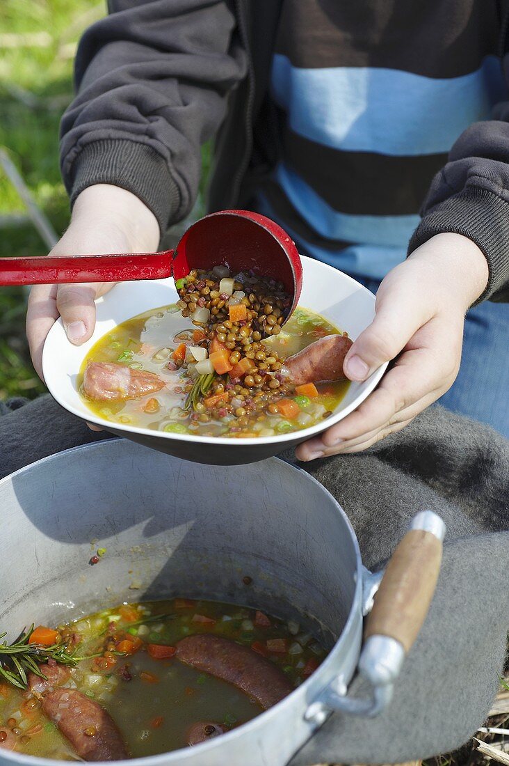 Ladling lentil and sausage stew into a dish at a picnic