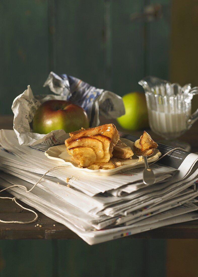 A piece of apple tart on newspapers with a jug of cream