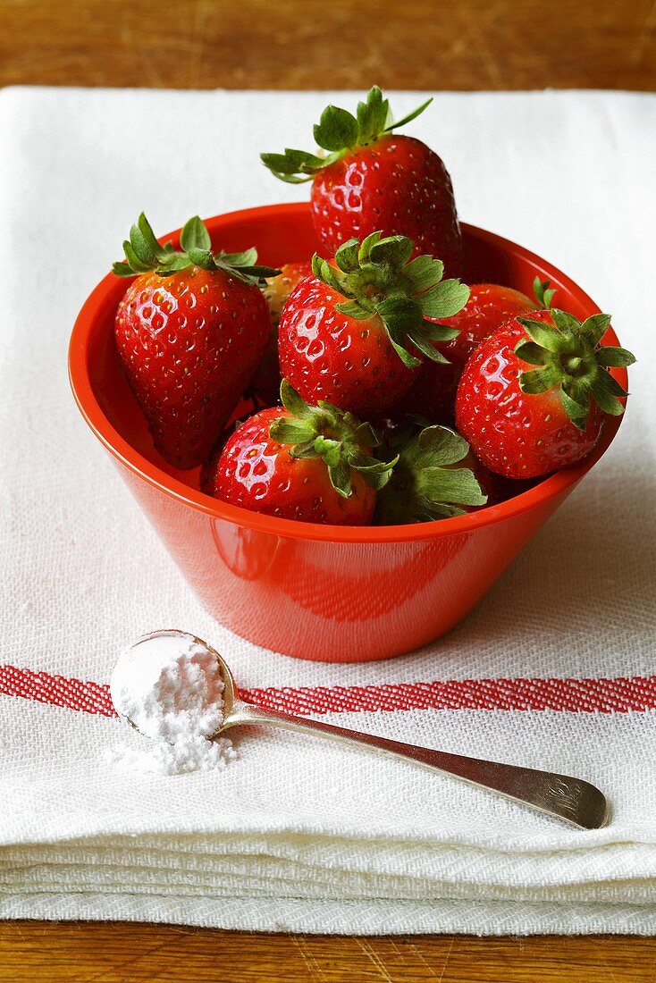 Fresh strawberries in red bowl, spoonful of sugar beside it