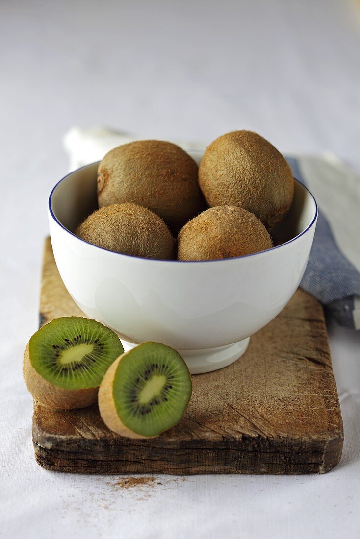Whole and halved kiwi fruit in and beside bowl on wooden board