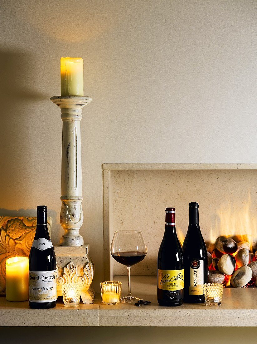 Rotweinflaschen, Rotweinglas und Kerzen vor dem Kamin