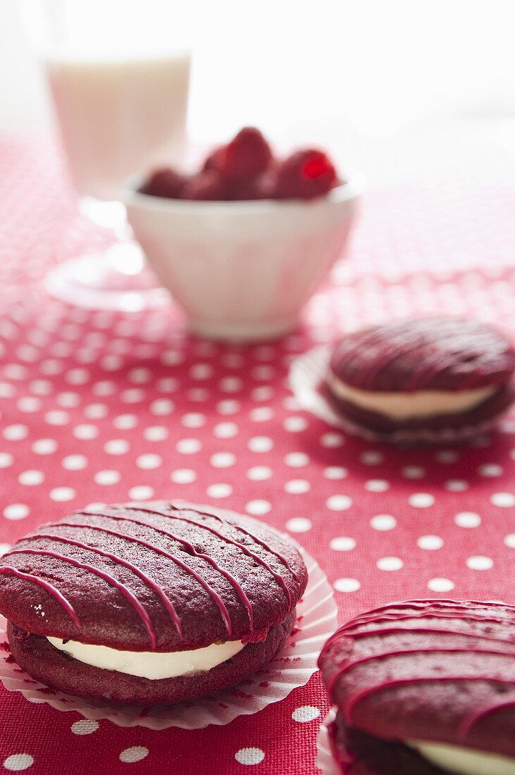 Raspberry whoopie pies on a spotted tablecloth with fresh raspberries and a glass of milk