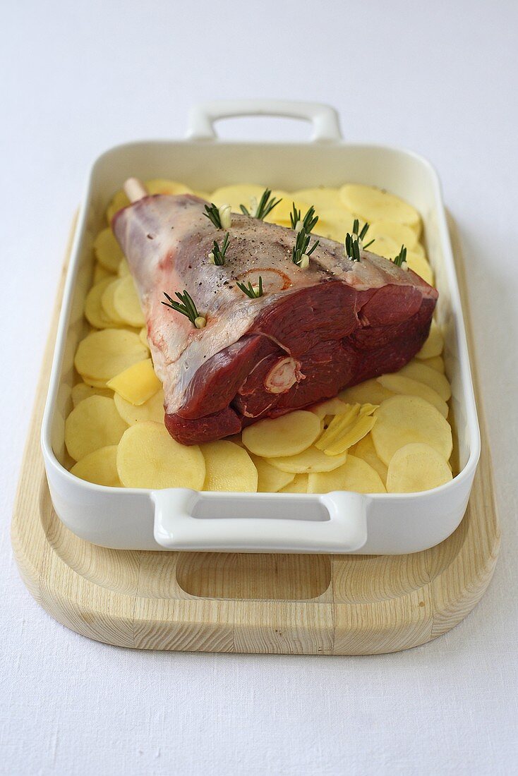 A leg of lamb stuck with rosemary on a bed of sliced potatoes (ready to roast)