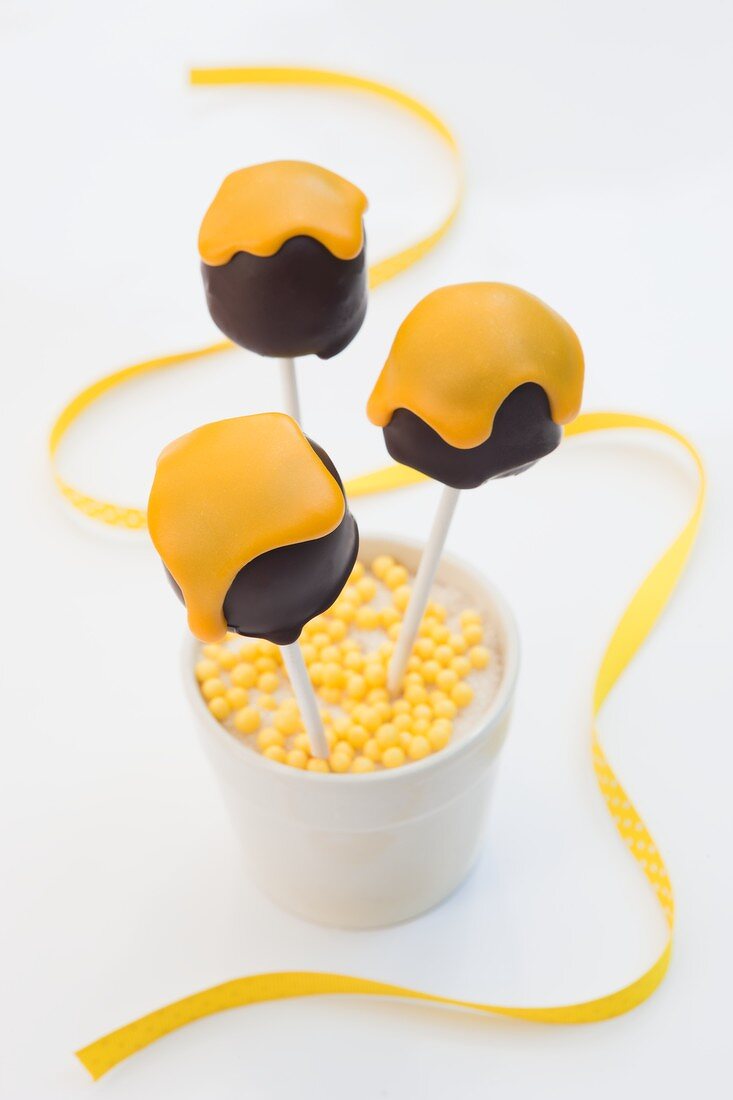 Cake pops shaped like chocolate marshmallows with yellow icing in a cup of yellow sugar beads