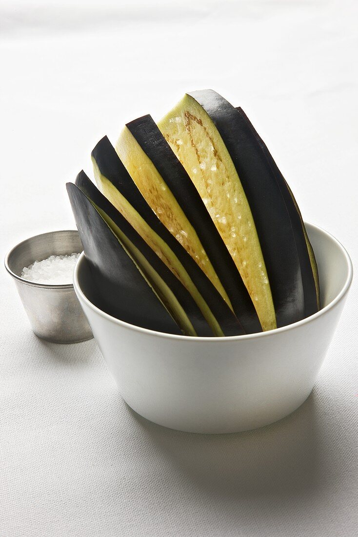 Aubergine slices in a bowl with a pot of salt