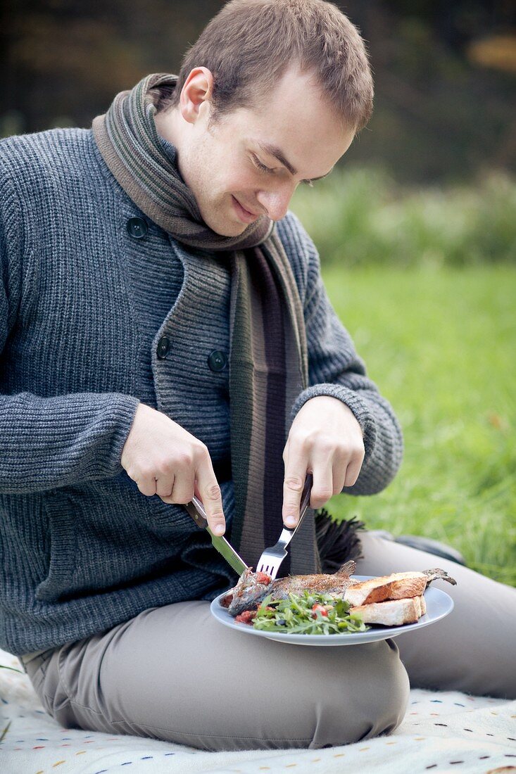 Mann sitzt auf Picknickdecke und isst Steckerlfisch