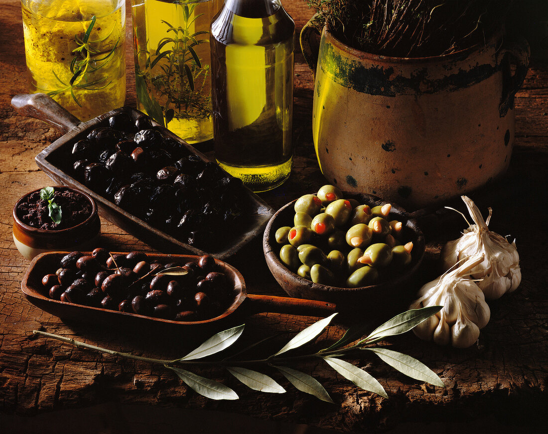 Mediterrane Produkte: verschiedene Oliven in Schalen, Knoblauch, Olivenöl und Kräuter