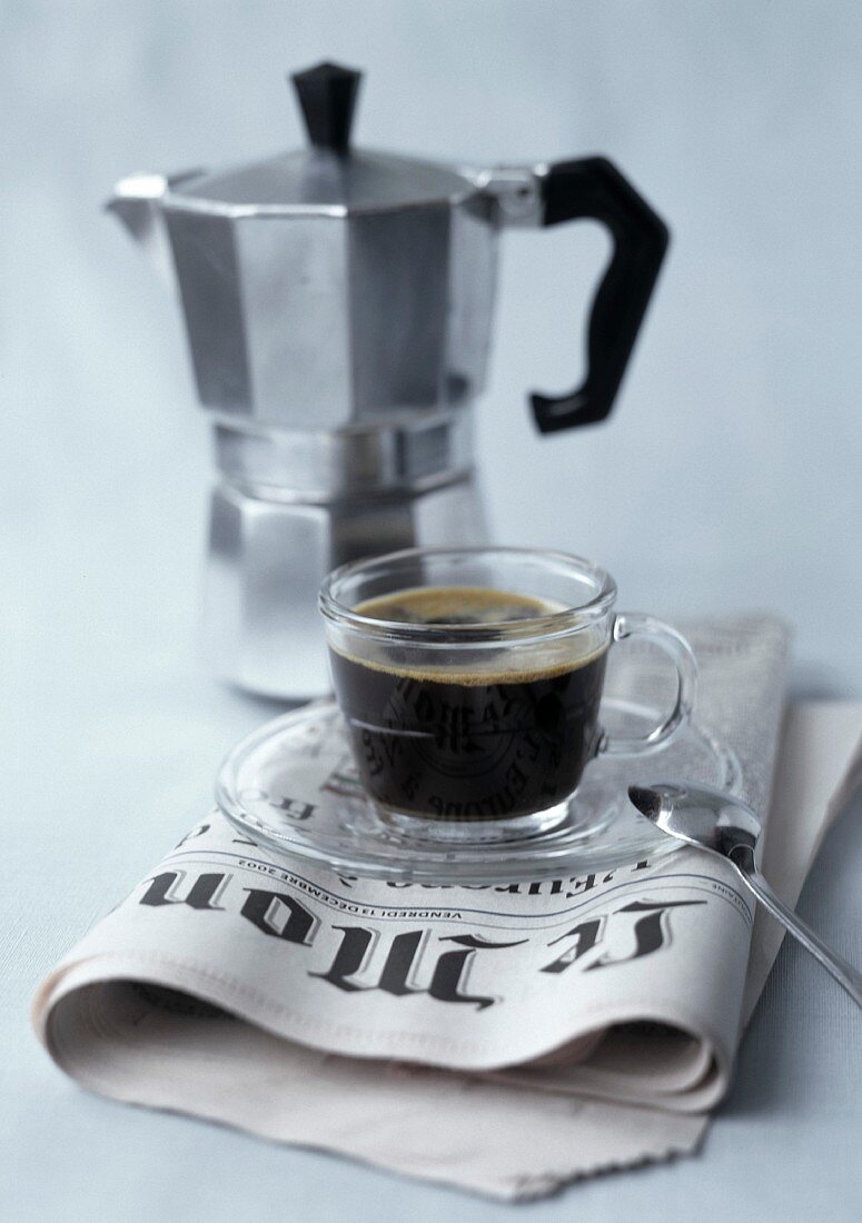 Glastasse mit Kaffee auf Zeitung, Kaffeekocher im Hintergrund
