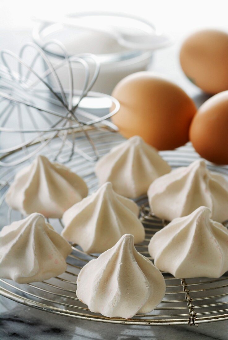 Baiserhauben auf einem Kuchengitter, im Hintergrund Eier, Schneebesen und Zucker