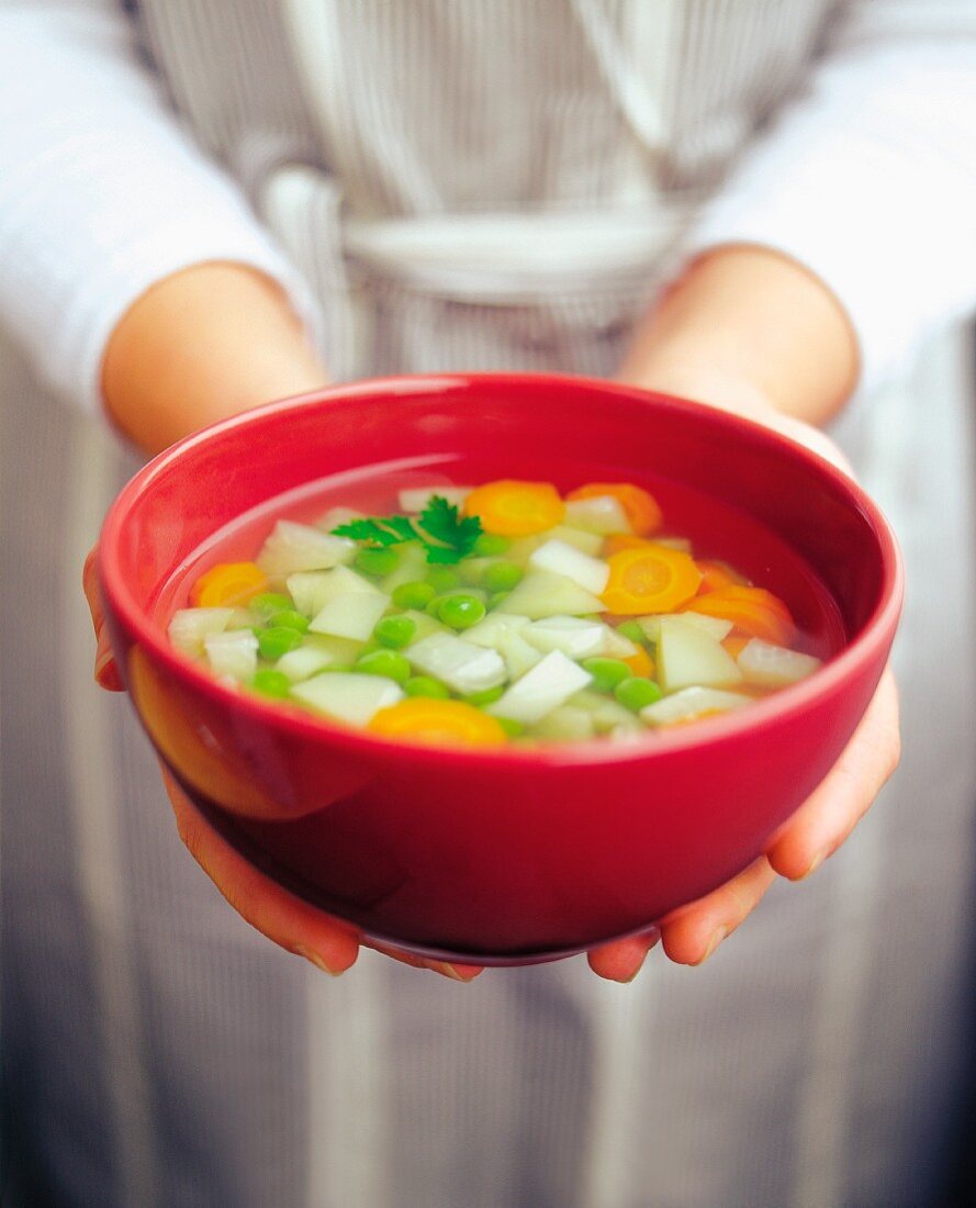 A woman holding a bowl of soup with potatoes, carrots and peas