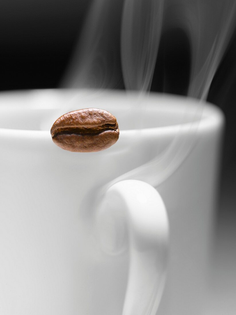Coffee bean on the edge of a steaming hot cup of coffee