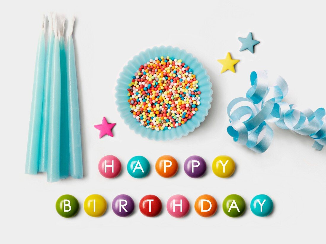 'Happy Birthday' written on Smarties and a blue paper cup full of sugar balls for decorating cakes, ribbon and blue birthday candles