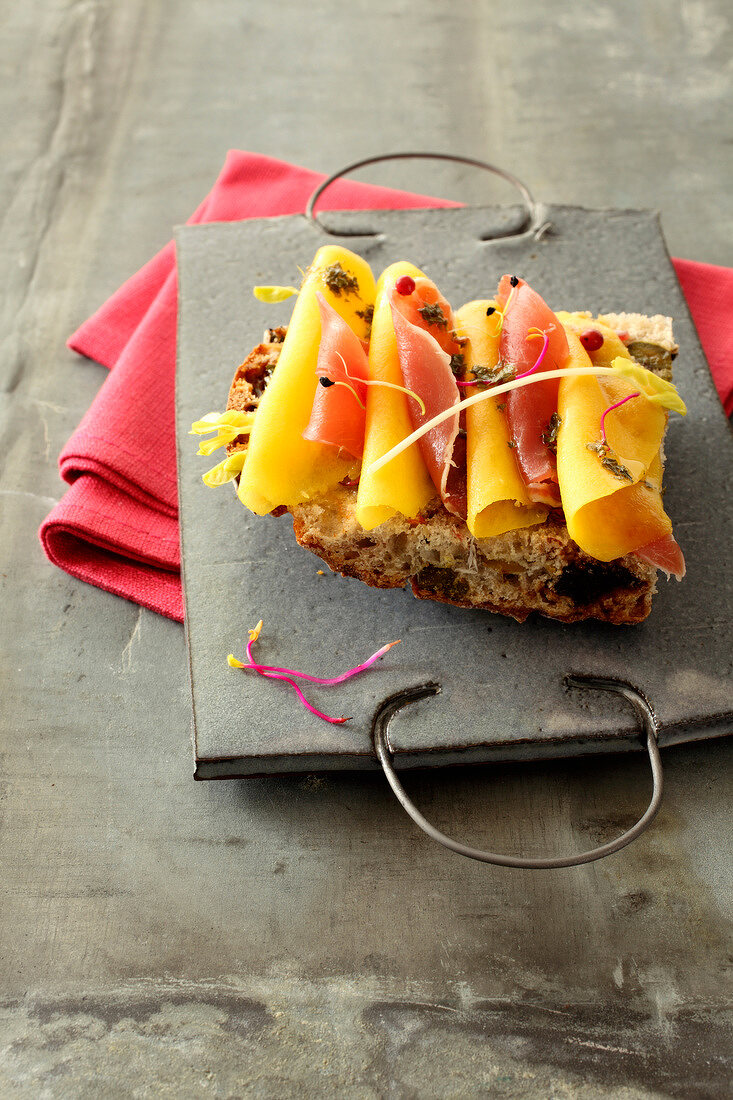 Hauchdünne Mango- und Rohschinkenscheiben mit Knoblauchsprossen und Petersilienöl auf Brot mit kandierten Früchten