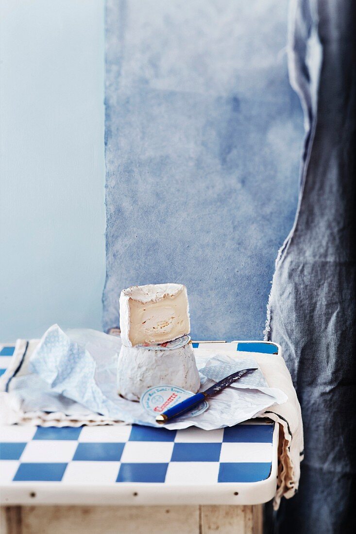 Weichkäse mit Käsemesser und Papier auf weiss-blau karierter Tischplatte