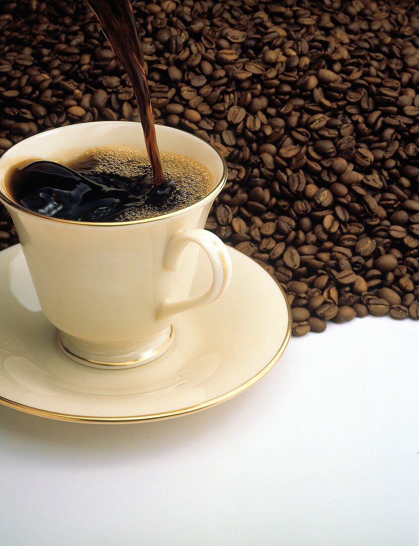 Kaffee wird in Tasse gegossen; Untergrund: Kaffeebohnen