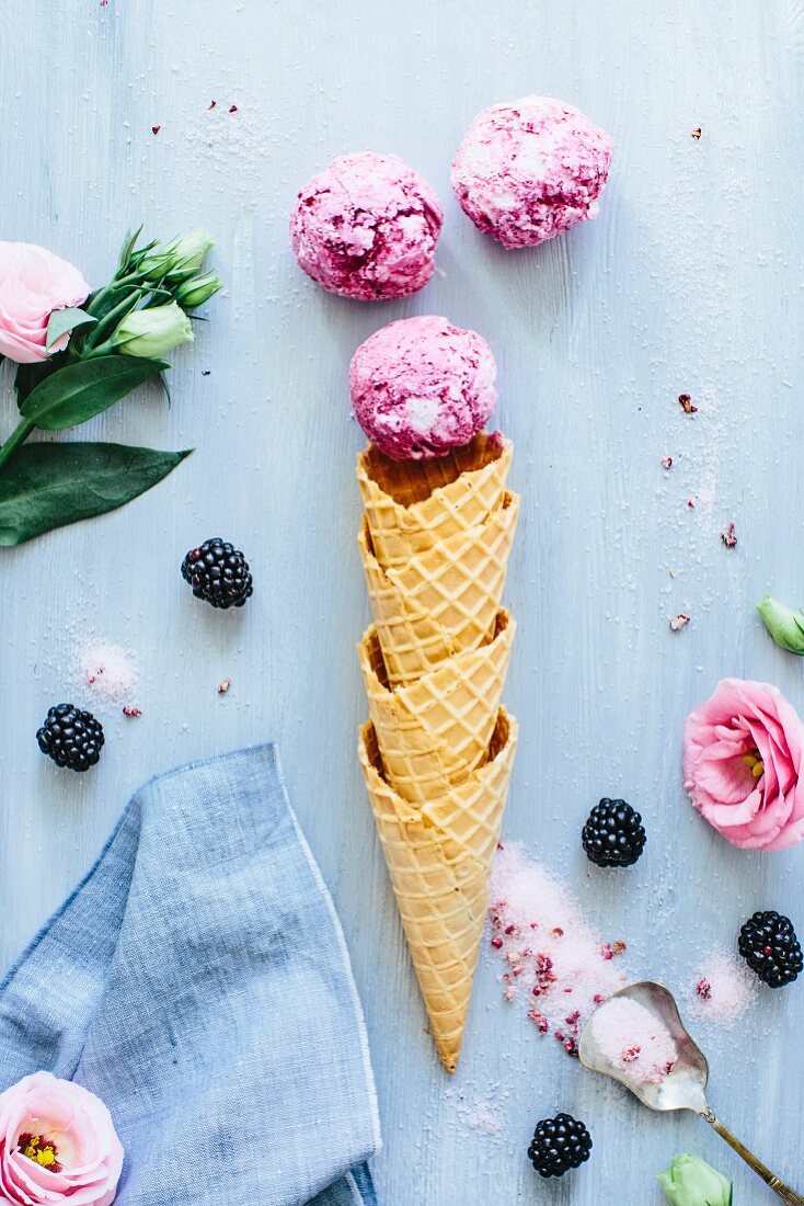 Komposition mit Eistüten, Kugeln Schokoladen-Brombeer-Eiscreme, Blüten und Löffel mit Zucker
