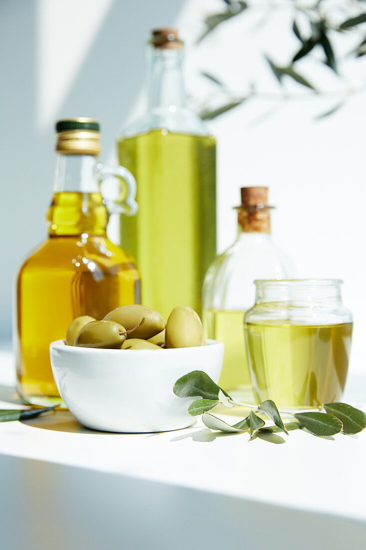 Various bottles of aromatic olive oil, jar, bowl with green olives and branches on white table