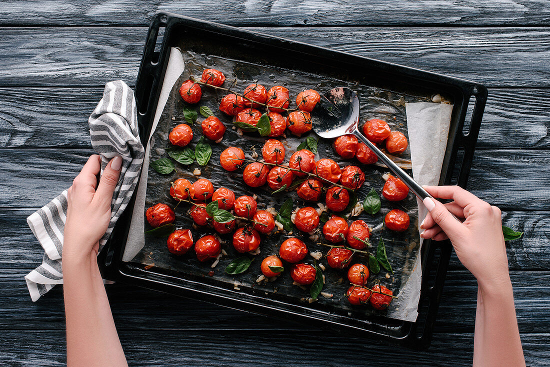 Woman pouring sauce on baked tomatoes in pan on dark wooden table