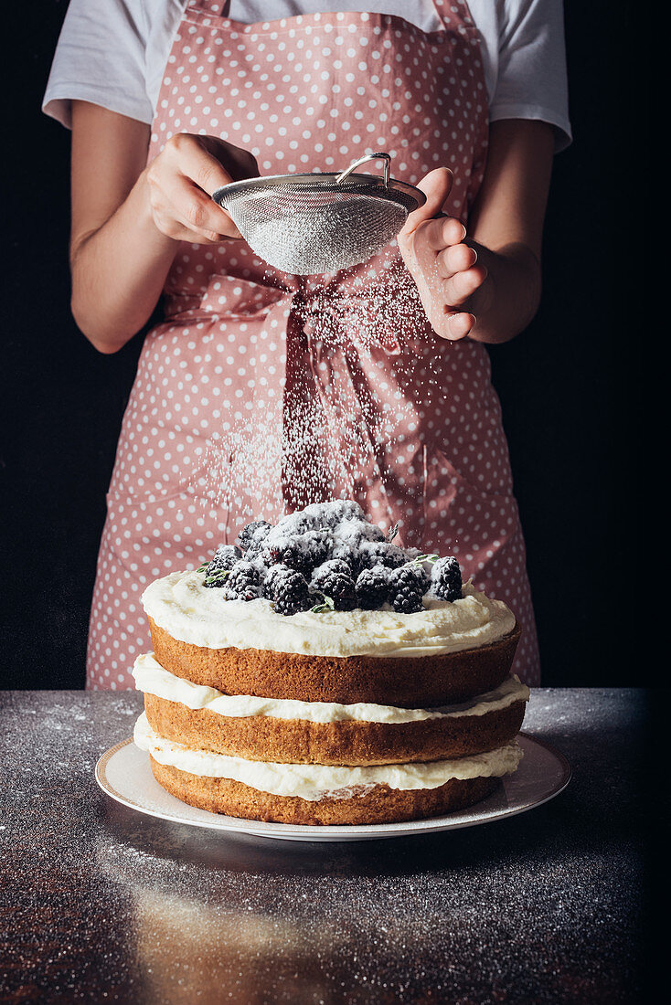 Frau siebt Puderzucker über Schichttorte mit Brombeeren