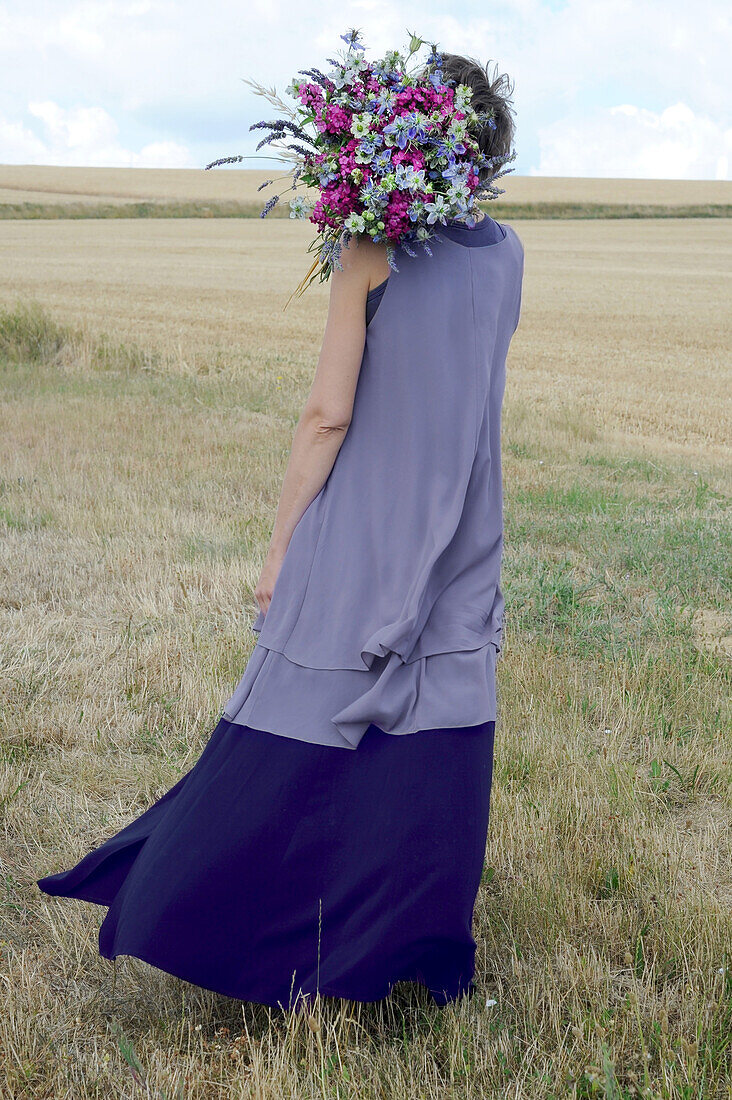 Back view of woman in purple dress standing in field with bouquet of flowers on shoulder