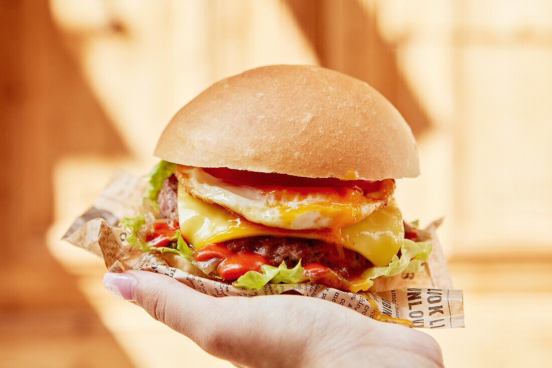 Hand holding a chicken burger with tomato, cheddar, egg, lettuce and ketchup