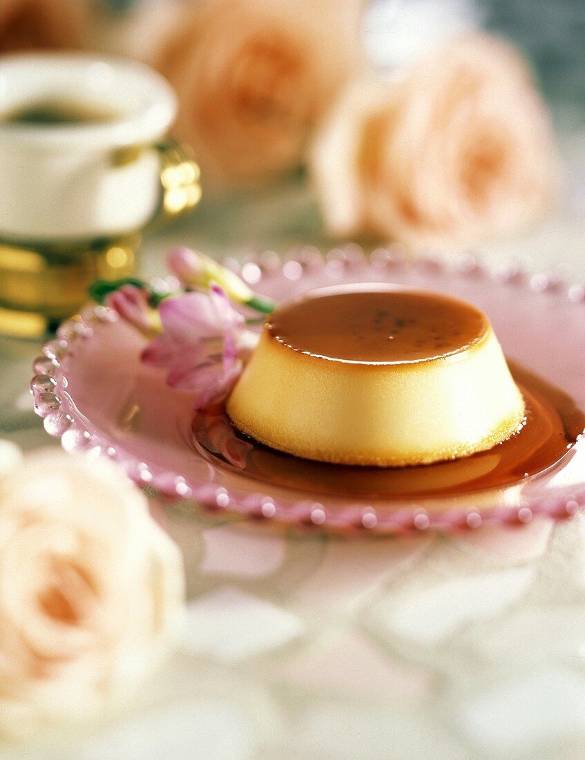 Karamellflan mit Sauce auf rosa Glasteller; Deko: Blumen