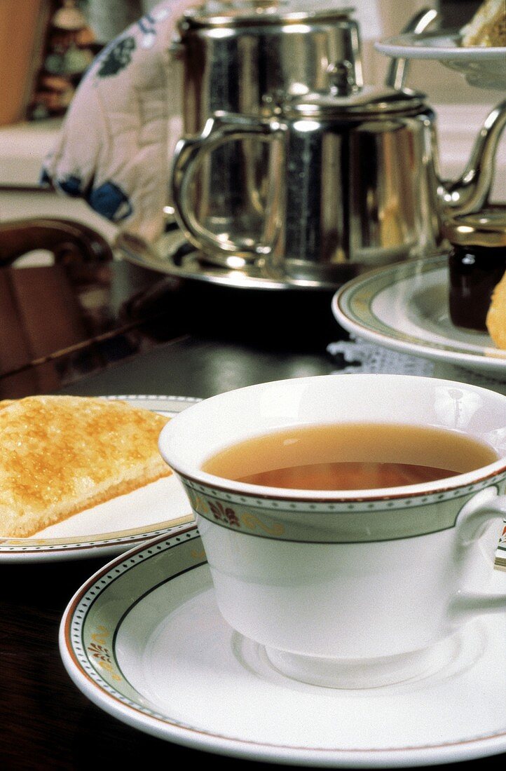 Tee in Tasse vor Teekannen, Kuchen etc. im Hintergrund