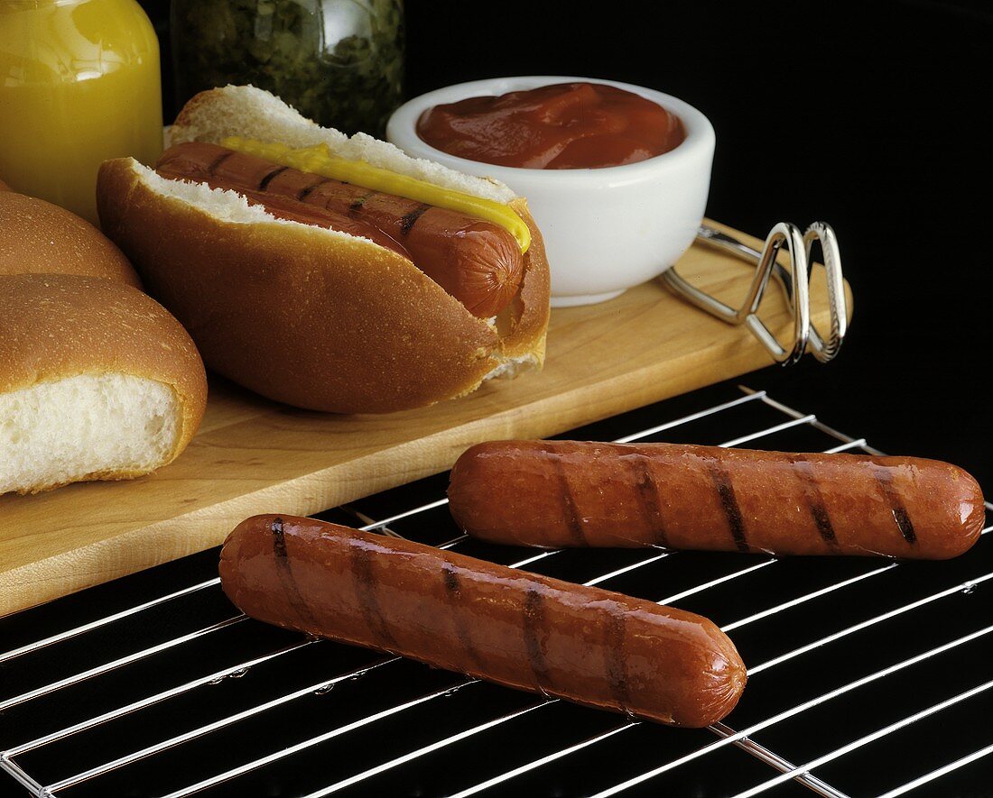 Hot Dog mit Senf und Ketchup, davor zwei Würstchen am Grill