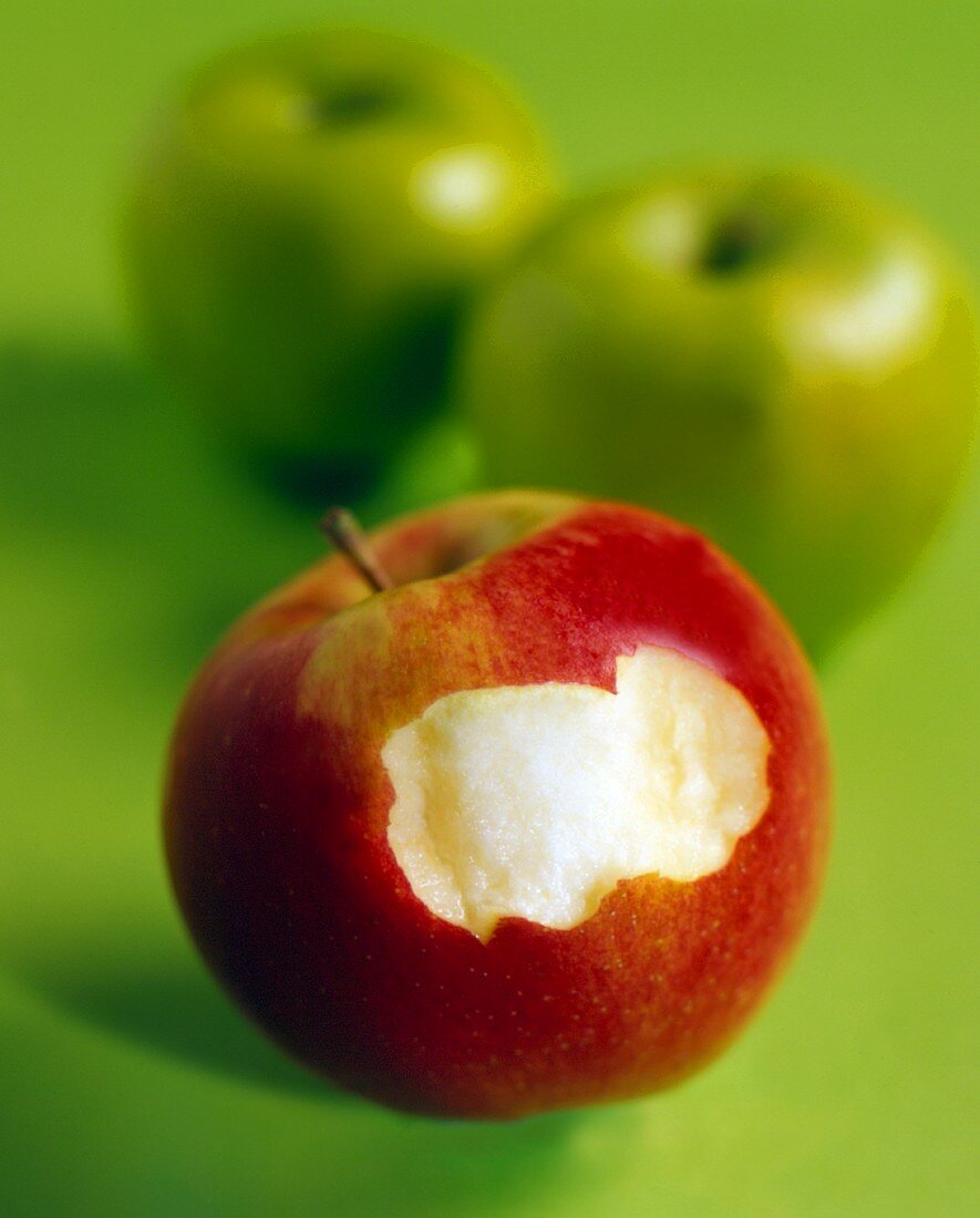Red apple, a bite taken, in front of two green apples