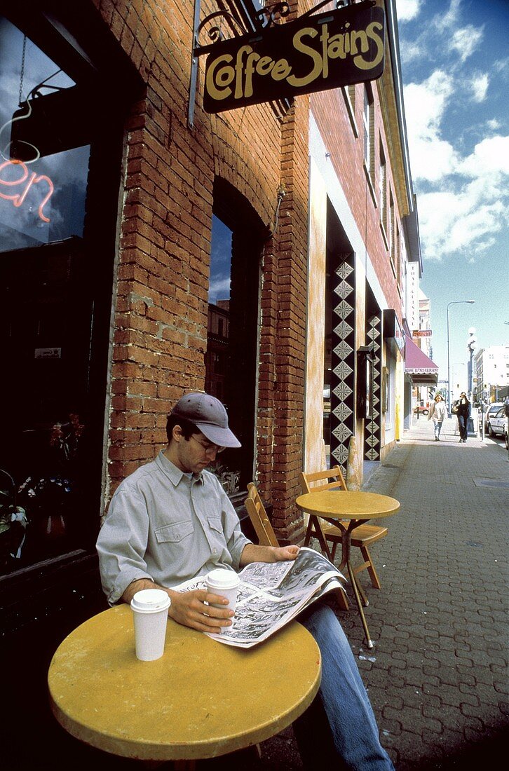 A Man Sitting Outside a Coffee Shop Reading a Newspaper