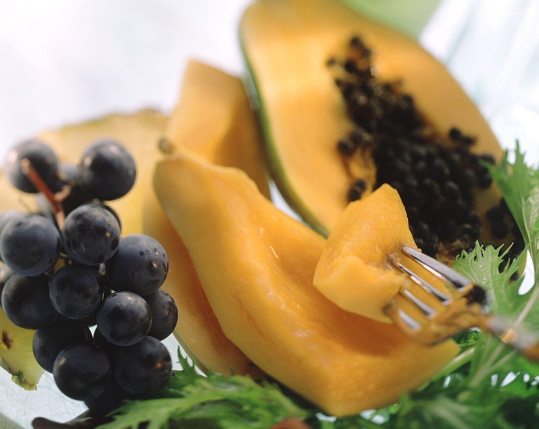 Aufgeschnittene Papaya mit Stück auf Gabel; rote Trauben