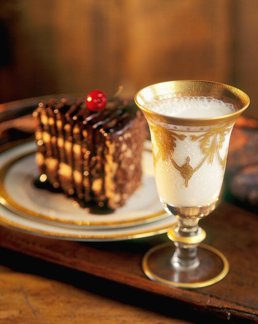 Milch in Glas mit Goldverzierung, im Hintergrund ein Stück Torte