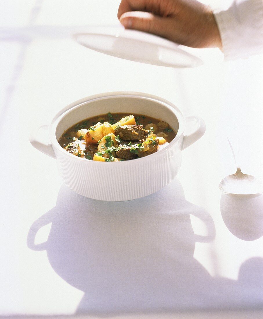 Meat stew in soup tureen, hand holding lid above it