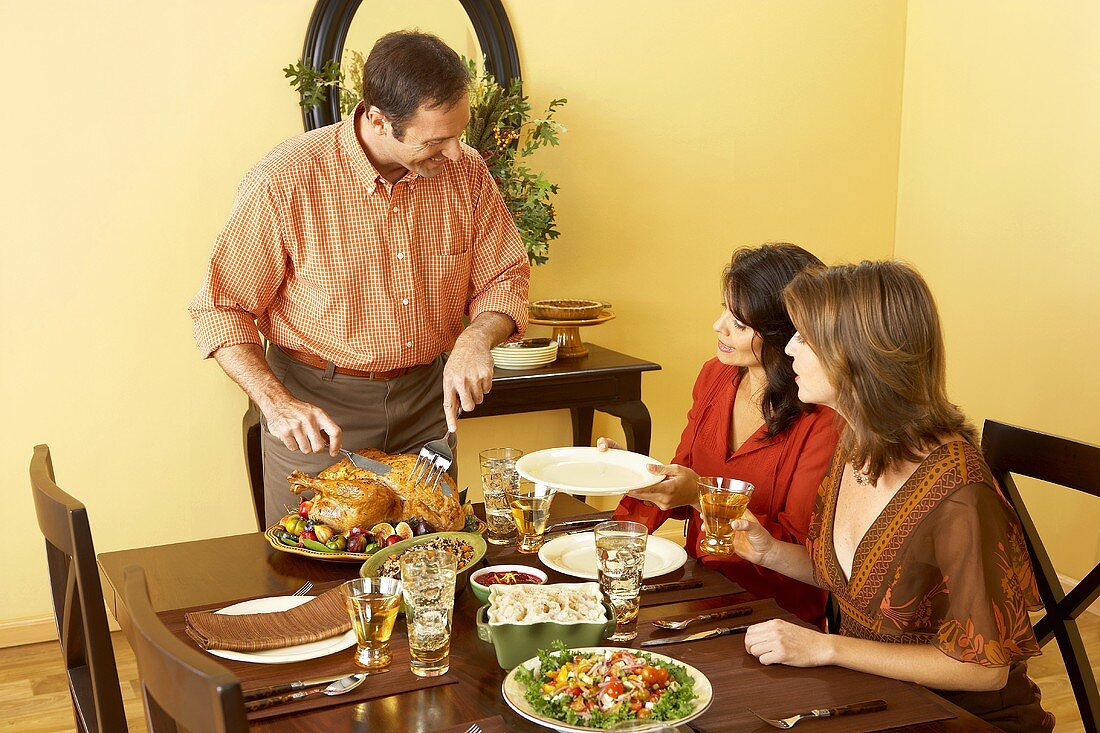 Man Carving Thanksgiving Turkey at Dining Table, Women Seated at Table